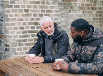 Two men sat on a bench having a coffee and talking